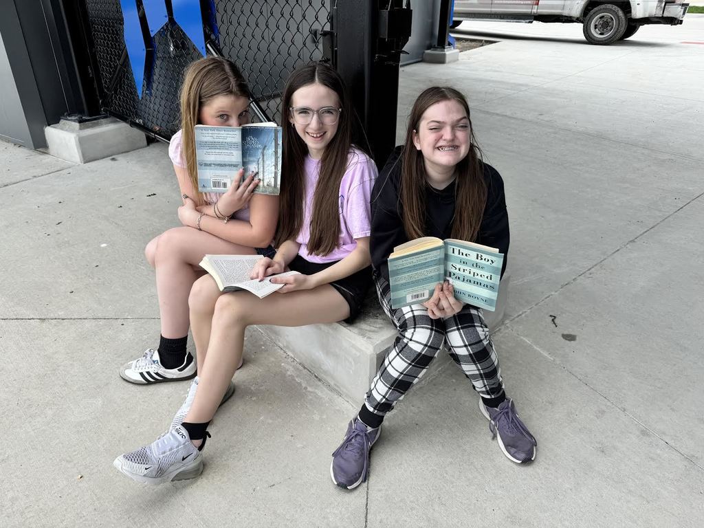 Three girls sitting on a concrete step outdoors, each holding and reading a book on a clear day.