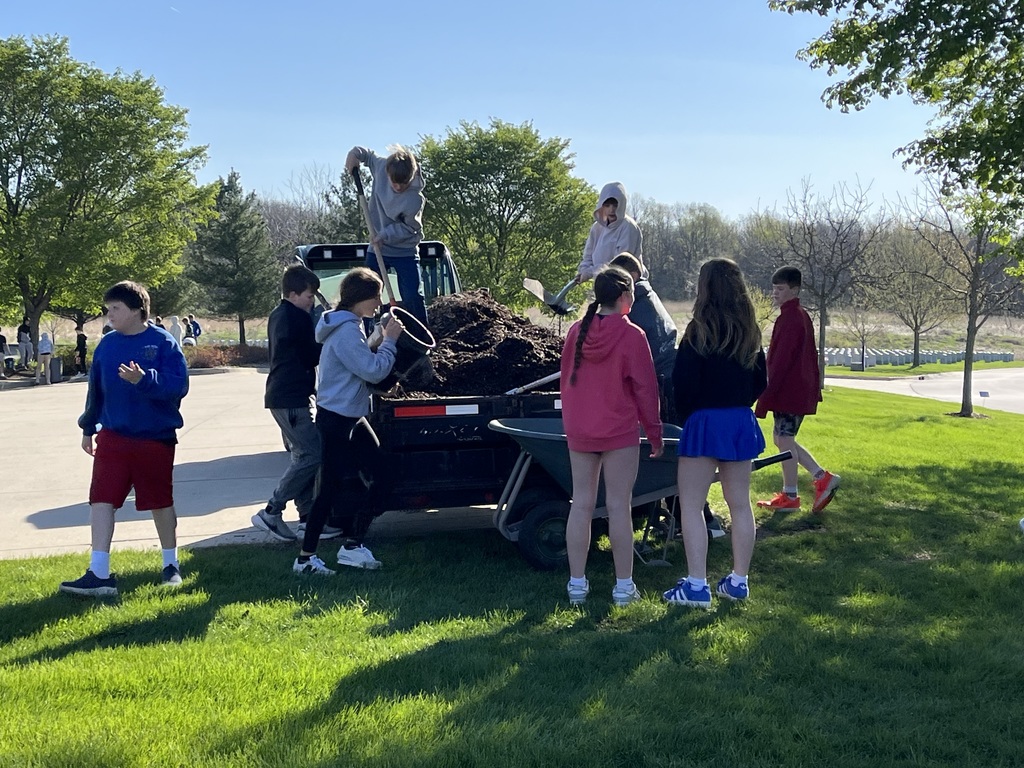 Group of students gather around a trailer, shoveling mulch into a wheelbarrow under bright sunlight in a park-like area.