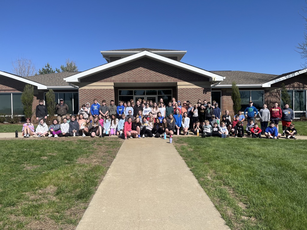 Large group of students and adults pose together outside a brick building, seated and standing along a walkway.
