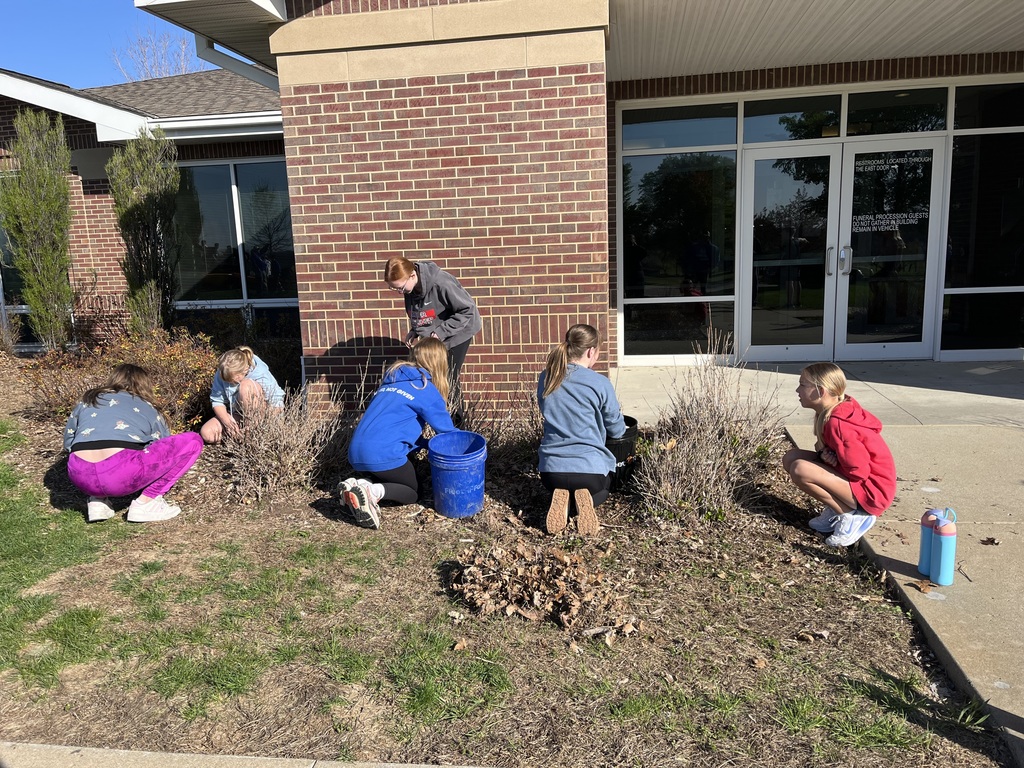 Children kneel in a garden bed by a brick building, pulling weeds and collecting debris in buckets on a sunny day.