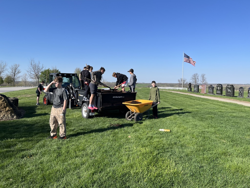 Boys shovel soil from a utility trailer into a yellow wheelbarrow on a grassy field near headstones and a U.S. flag.