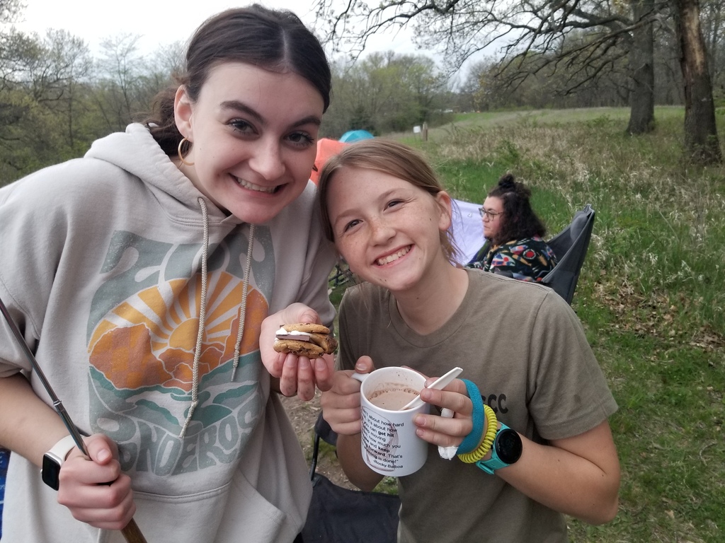 Two girls smile while holding a s’more and a mug of hot chocolate, standing at a campsite with chairs and tents behind.