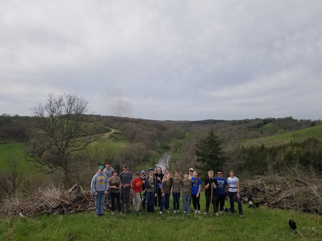 Large group poses on a hilltop overlooking a winding river and wooded valley, with piles of branches on either side.