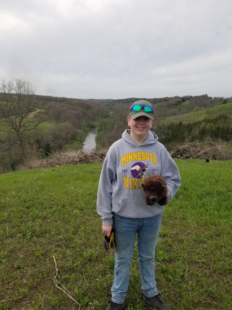 Person in a Minnesota Vikings hoodie stands on a grassy hill holding gloves and a small plush toy, river valley behind.