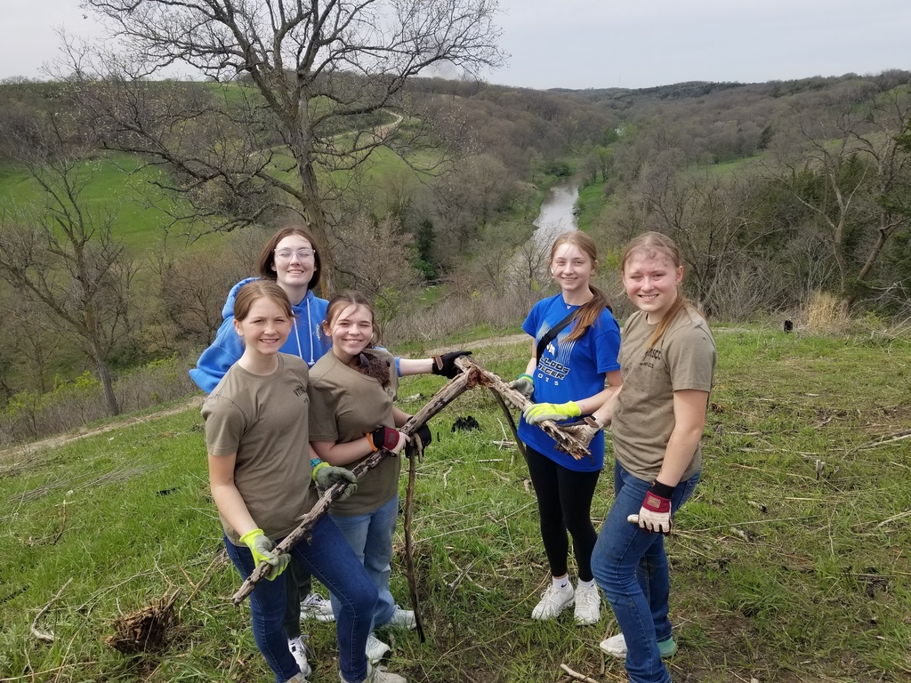 Five girls stand together on a hillside holding branches, smiling, with a river valley and trees in the background.