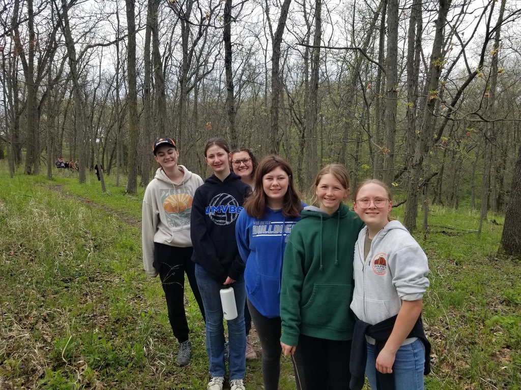 Six girls stand on a wooded trail, smiling at the camera, with leafless trees and green undergrowth around them.
