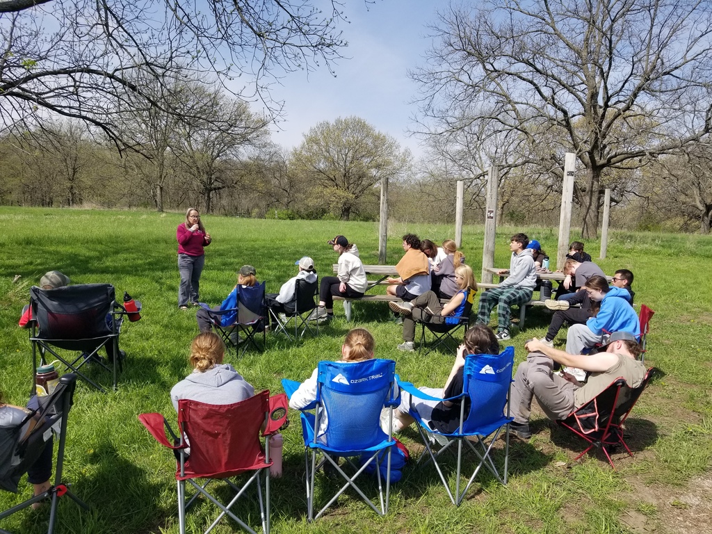 Group of teens sit in folding chairs outdoors, listening to a speaker in a grassy clearing with trees and wooden posts.