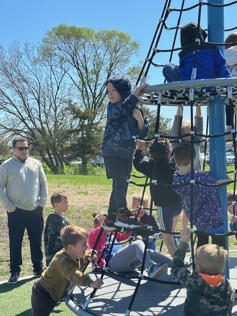Several children climb and play on a rope playground structure outdoors while an adult stands nearby watching.