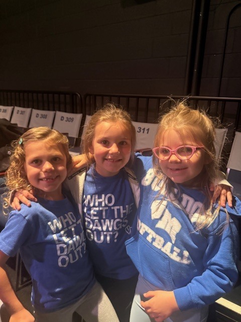 Three young girls stand close together, smiling with arms around each other, wearing matching blue “Who Let the Dawgs Out?” shirts.