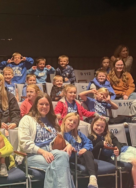 Children and a few adults sit together in theater seats, some making playful faces and peace signs, wearing blue shirts.