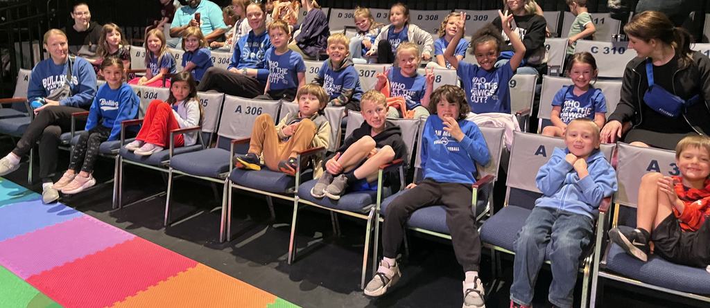 Group of young children and adults seated in rows, most in matching blue shirts, smiling and posing inside a theater.