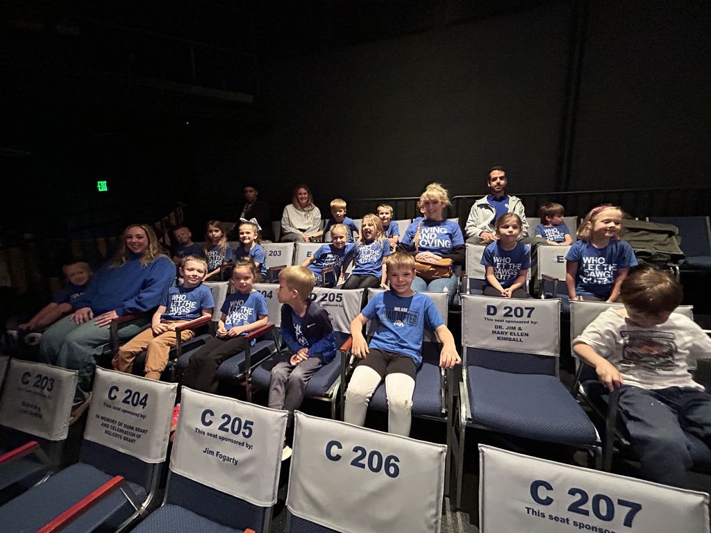Children and a few adults sit in theater seats, many wearing blue shirts, smiling and chatting before a performance.
