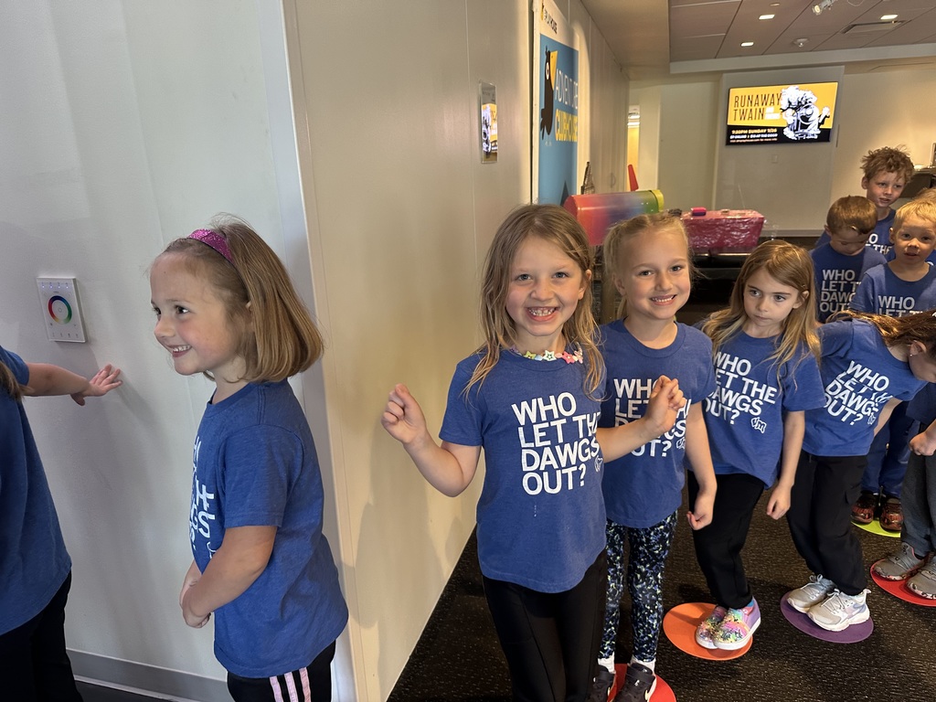Young children line up indoors on colored floor spots, smiling and wearing matching blue shirts, waiting their turn.