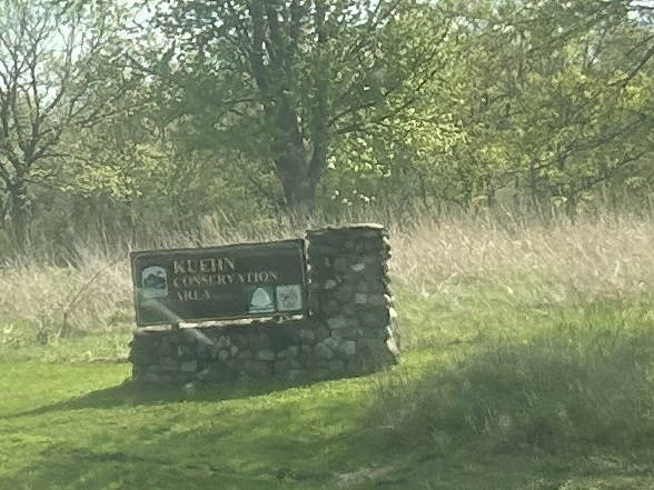 Stone sign reading “Kuehn Conservation Area” set in grass, with trees and tall prairie plants behind.
