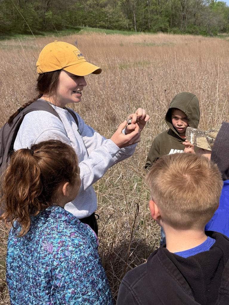 Educator in a yellow cap shows a small object to children gathered in a dry prairie field.