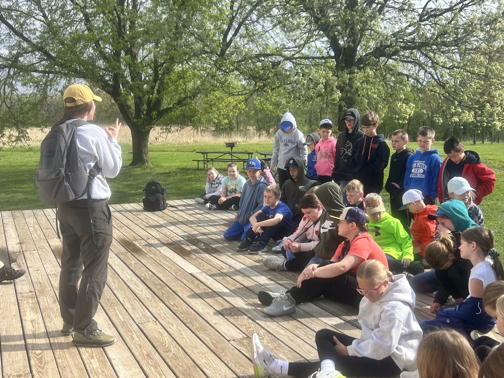 Instructor in a yellow cap speaks to seated children on a wooden deck under leafy trees.