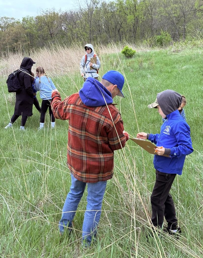 Students measure tall grasses with clipboards in a field while an instructor guides them.
