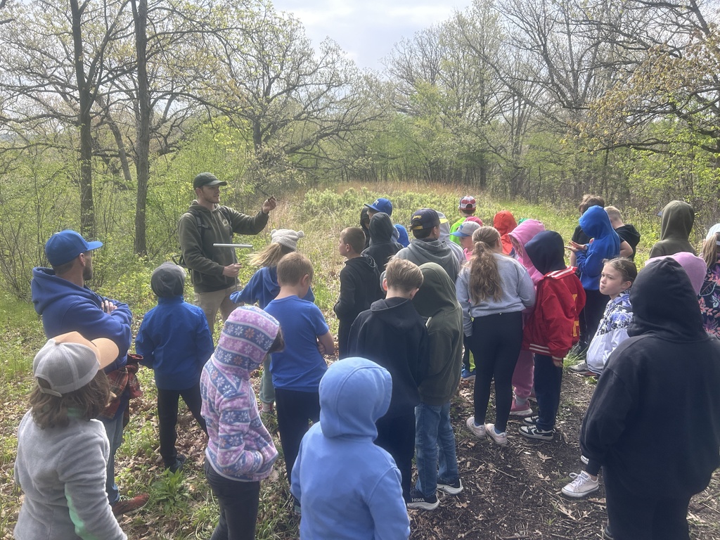 Naturalist addresses a group of children on a wooded trail, holding a tool while they watch.