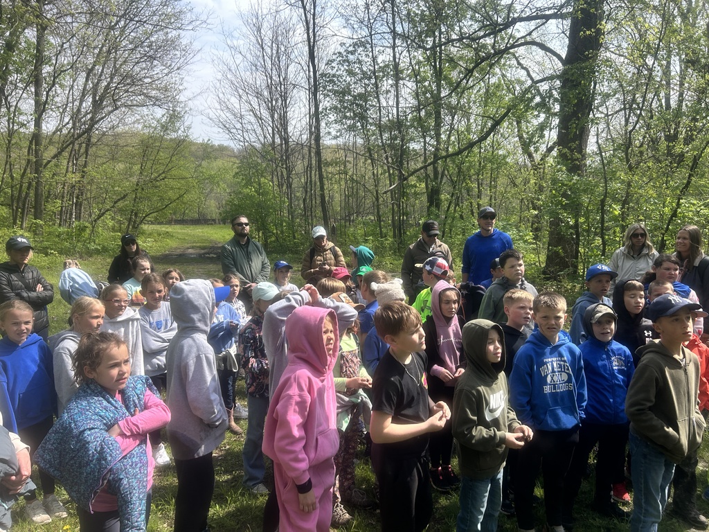 Large group of children and adults stand in a forest clearing, listening attentively outdoors.