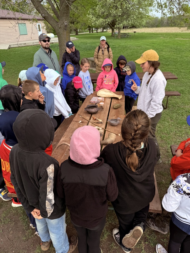 Children gather around a picnic table examining artifacts like bones and tools during a lesson.