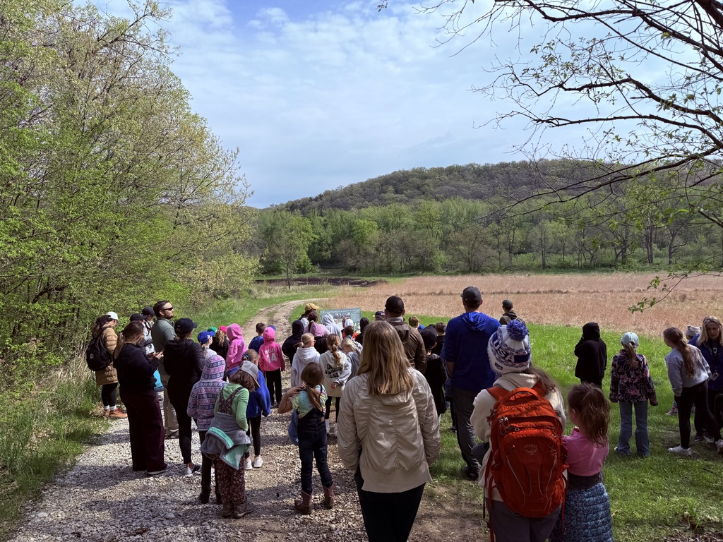 Group stands on a trail overlooking prairie and wooded hills, listening to a guide outdoors.