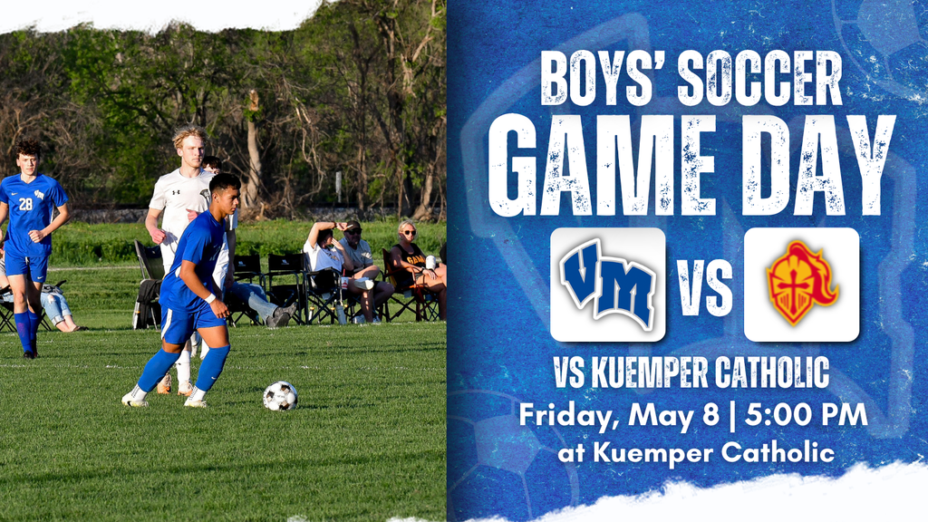 High school boys' soccer players in blue uniforms on the field during a game, with spectators seated in the background.