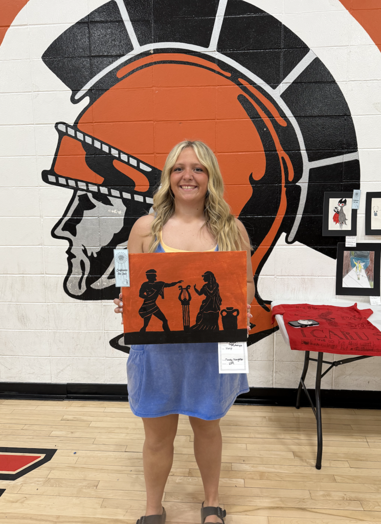 Young woman holding a painting of two ancient Greek figures in black silhouette against an orange background, standing in front of a large Trojan helmet mural.
