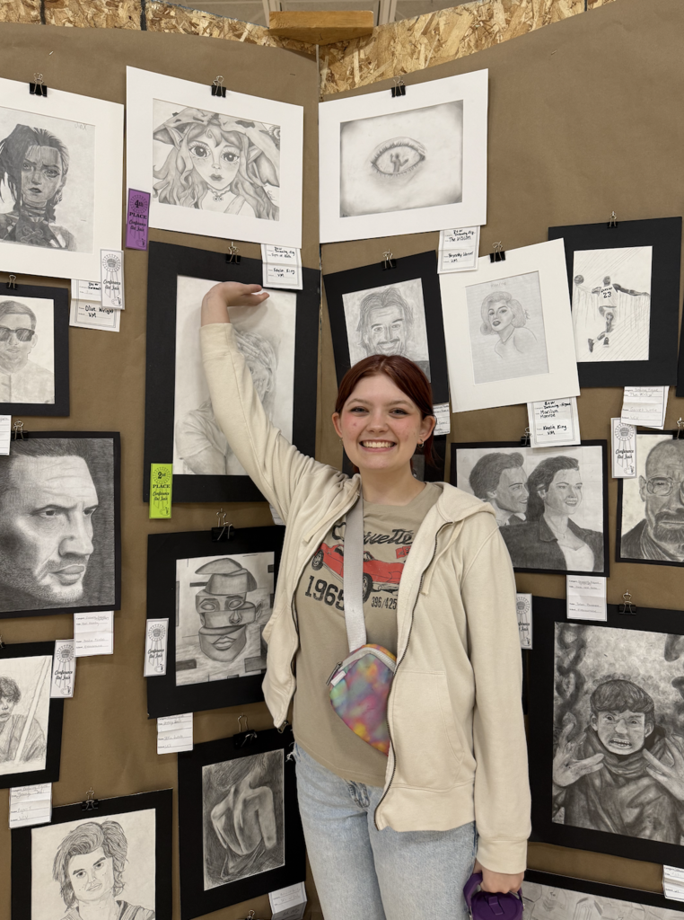 Person standing in front of a wall displaying various pencil sketches and charcoal portraits clipped to brown paper panels.