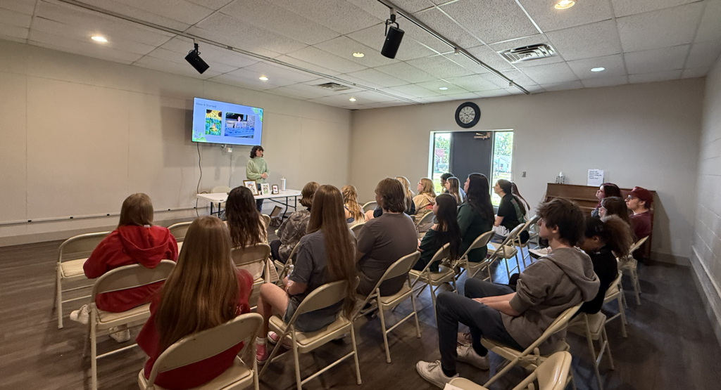 A group of people seated on folding chairs attentively watching a presenter explaining content displayed on a wall-mounted screen in a small conference room.