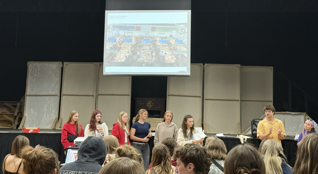 Group of young people standing on stage presenting to an audience with a projected image behind them.