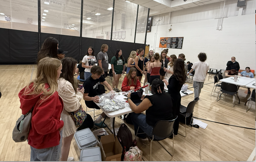 Students gather around tables in a gymnasium, engaging in an organized group activity or event with some seated and others standing in line.