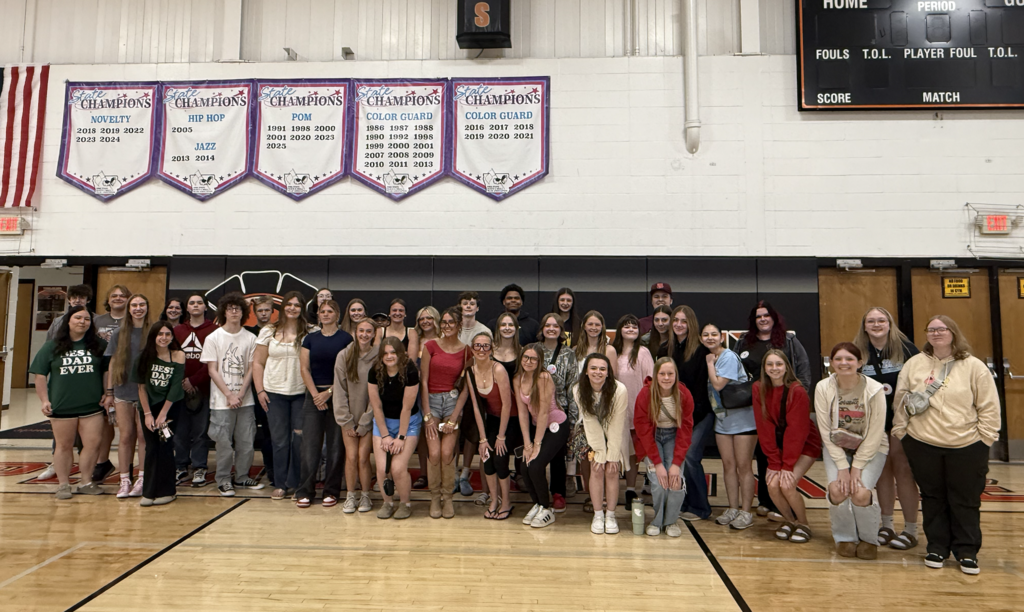 Large group of diverse young adults posing together in a gymnasium with championship banners hanging on the wall behind them.