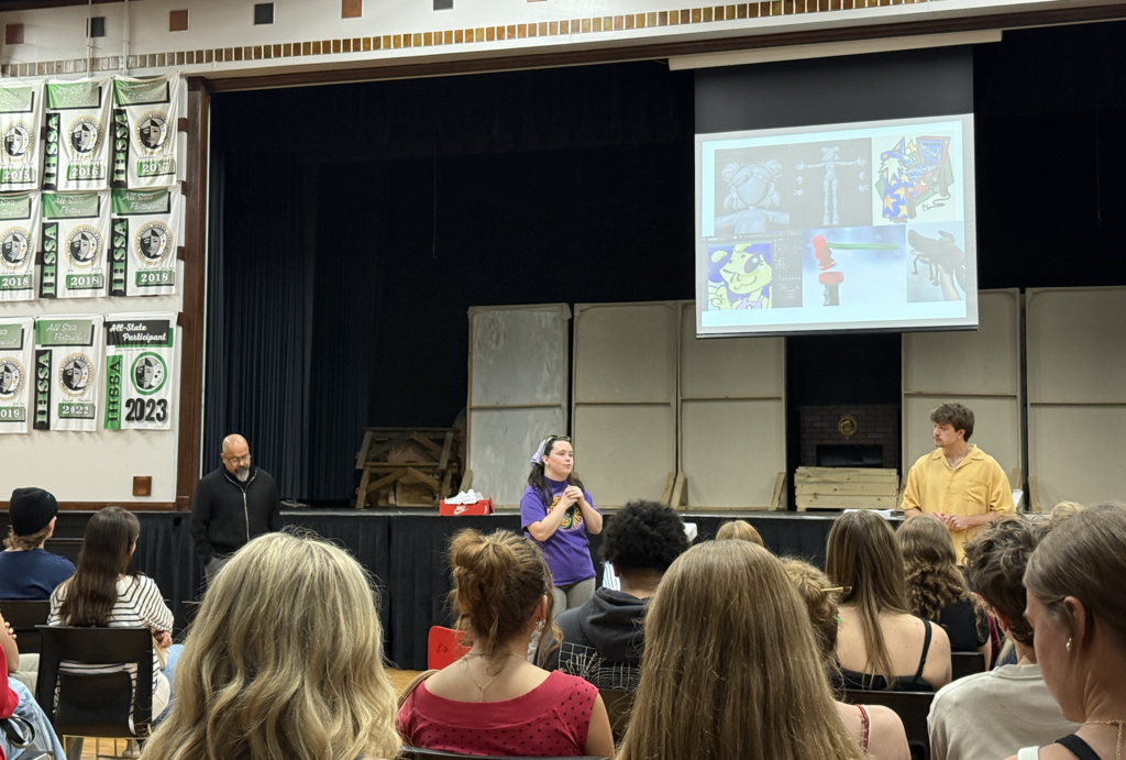 A speaker in purple addresses an audience in a school auditorium with two other presenters on stage and a multimedia presentation projected behind them.