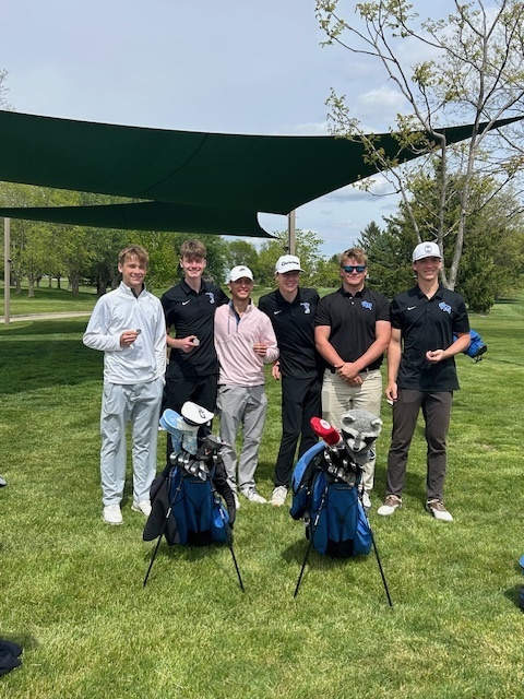 Group of six young men standing on a golf course with two golf bags in front of them under a shaded canopy.