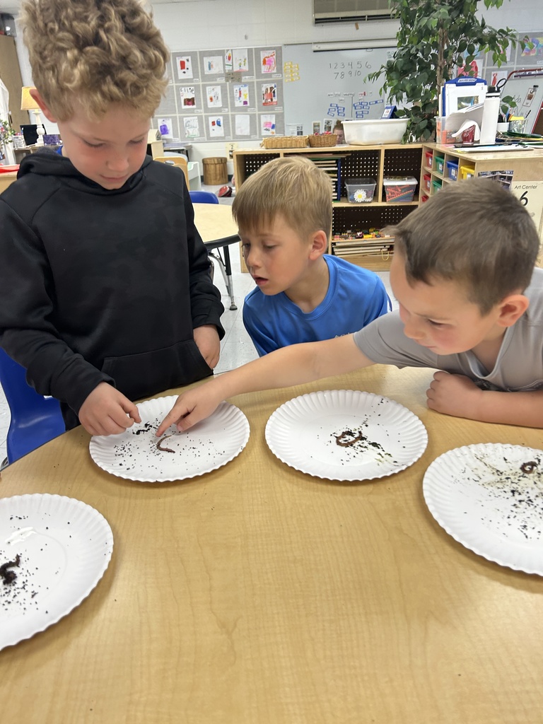Three children observe and point at worms on paper plates during a classroom science activity.