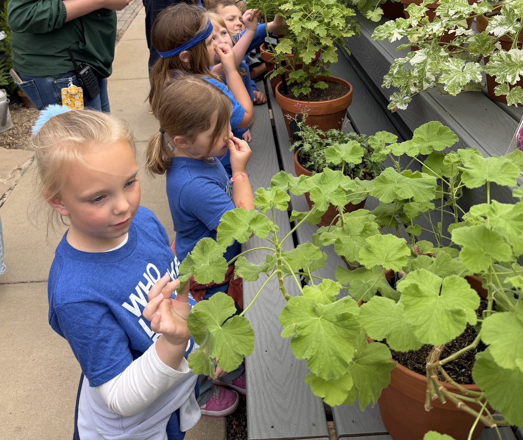 Children in matching blue shirts smell and examine potted plants during a greenhouse field trip.