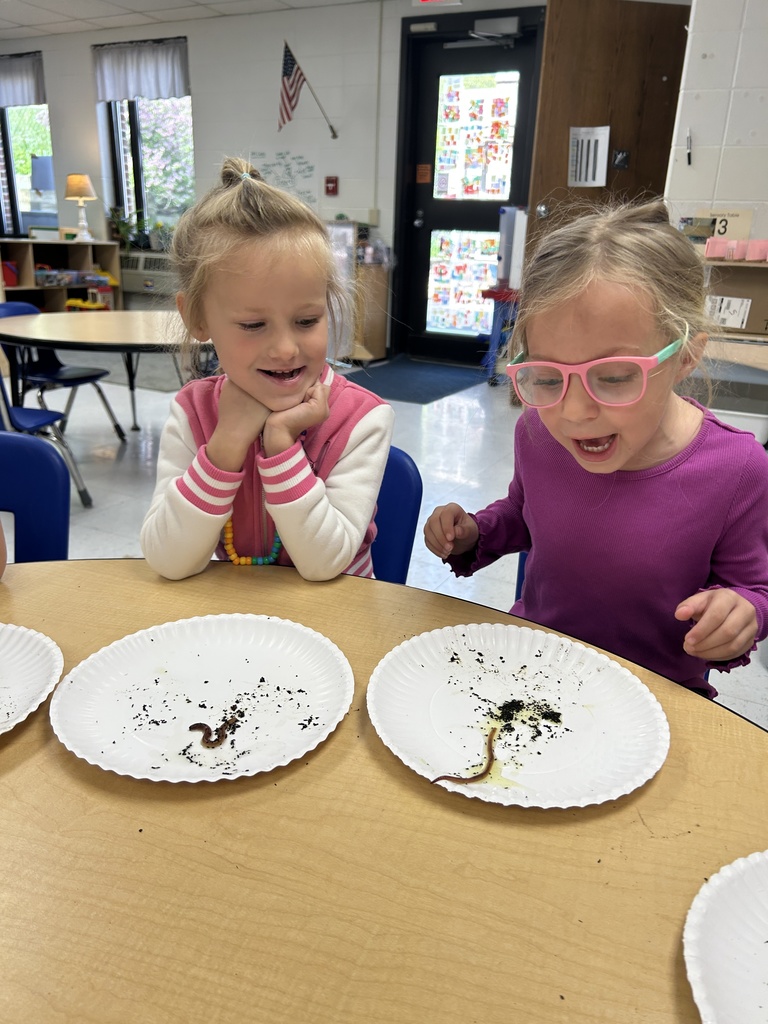 Two young girls smile excitedly while watching worms move across paper plates in a classroom.