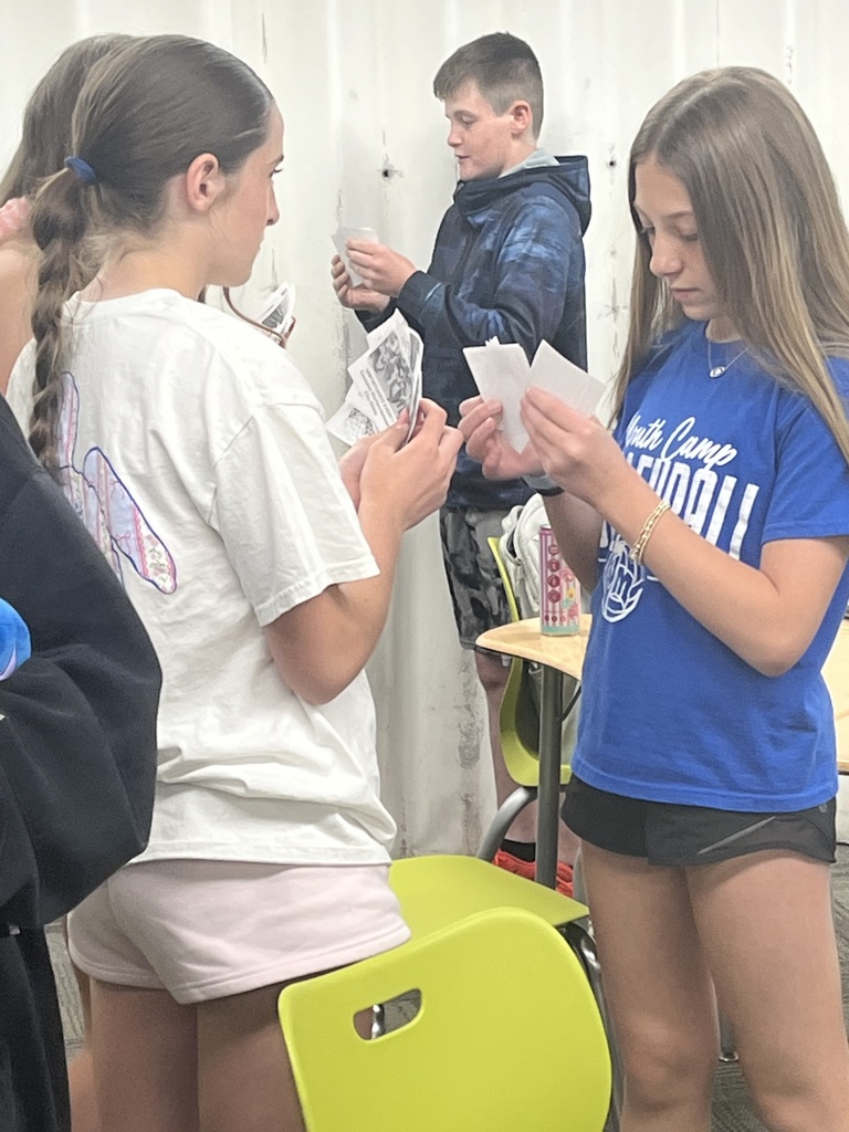 Three students stand in a classroom comparing and reading cards during an interactive learning game.