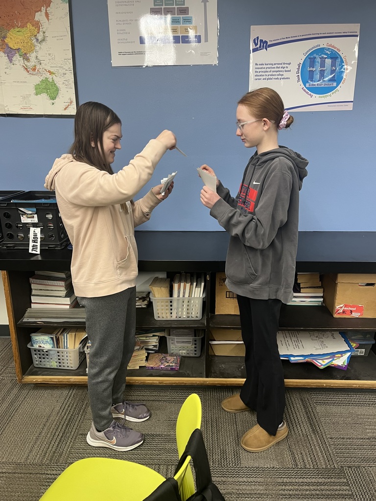 Two students smile while exchanging printed cards in a classroom activity beside bookshelves and maps.