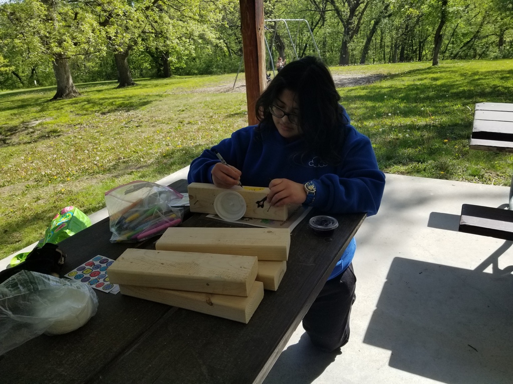 Student in a blue hoodie decorates wooden blocks with markers at a picnic table in a sunny park.