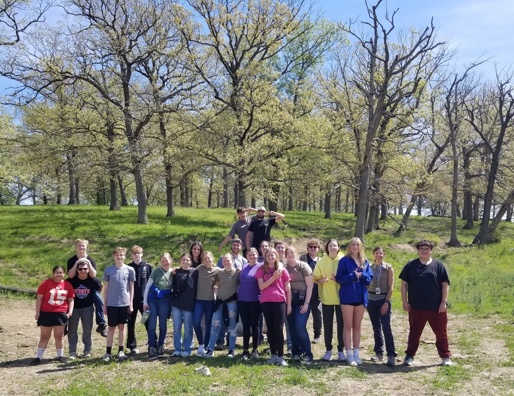 Students gather outdoors for a group photo in a sunny clearing surrounded by tall trees.