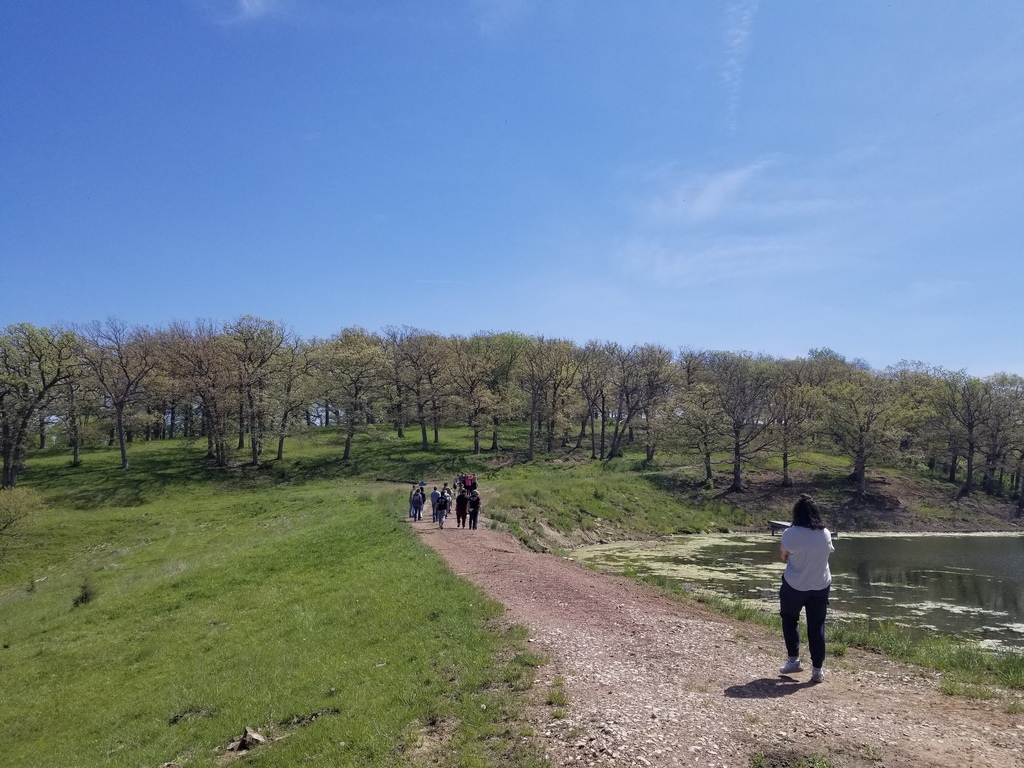 Students walk along a gravel path beside a pond surrounded by grassy hills and leafy trees.