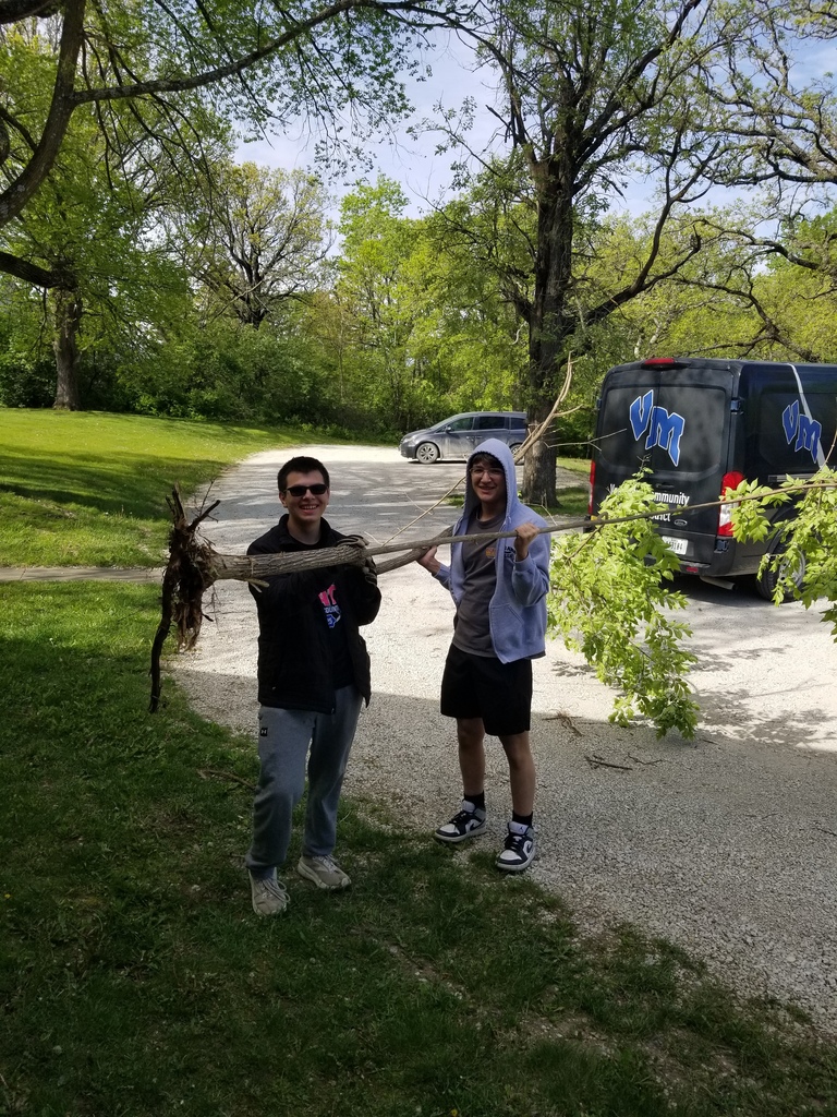 Two students smile while carrying a large fallen tree branch near a wooded gravel driveway.