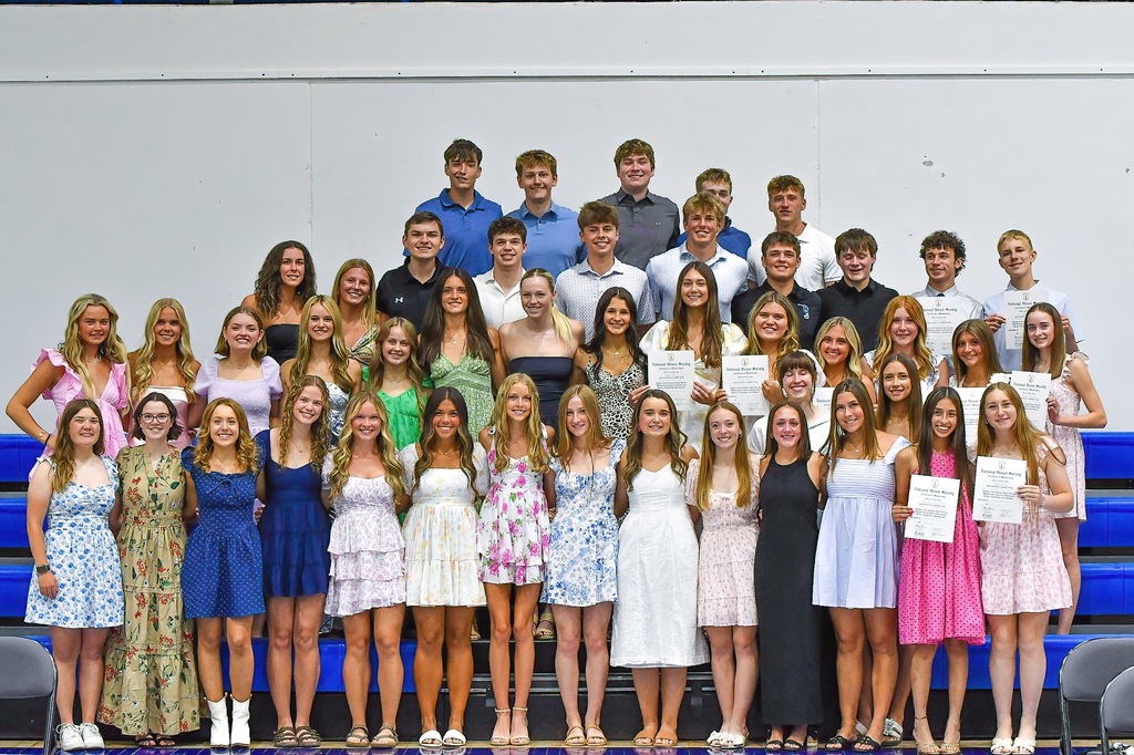 National Honor Society students gather for a large group photo on gym bleachers, several holding certificates.