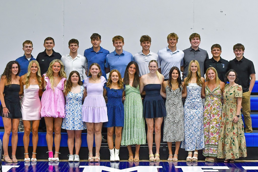 Large group of teenage students pose together in a gymnasium, dressed in colorful dresses and collared shirts.