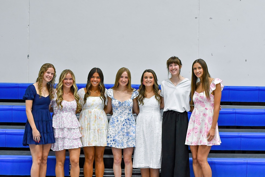 Seven young women stand arm in arm in front of blue gym bleachers, smiling and wearing pastel floral and solid-color dresses.