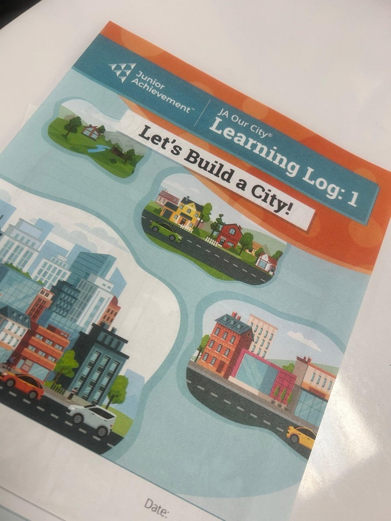 Cover of Junior Achievement “JA Our City” learning log titled “Let’s Build a City!” with illustrated neighborhoods and city scenes.