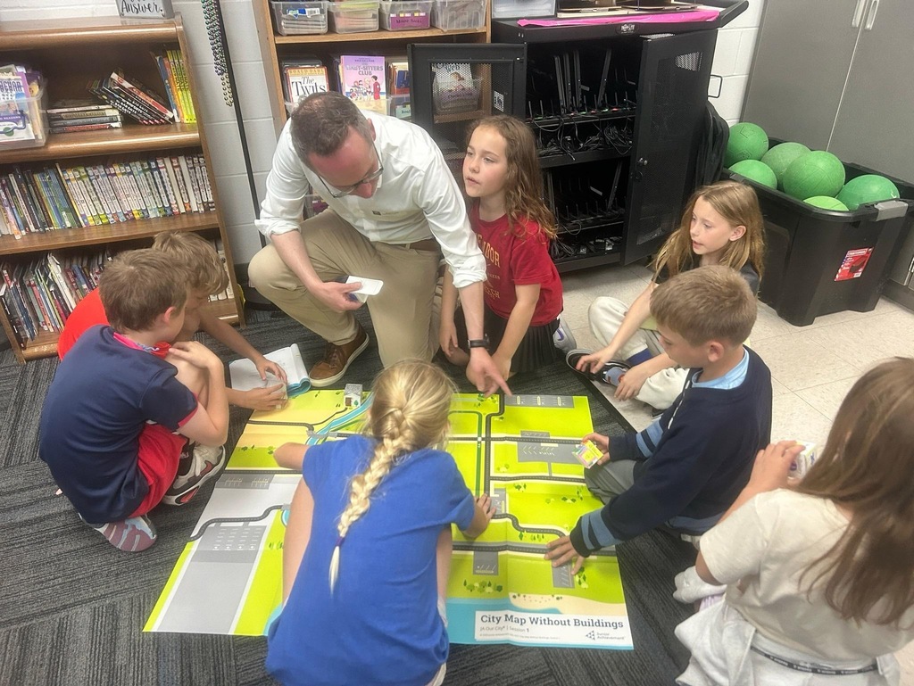 Adult volunteer and students gather around a city map, discussing placement of buildings during a hands-on activity.