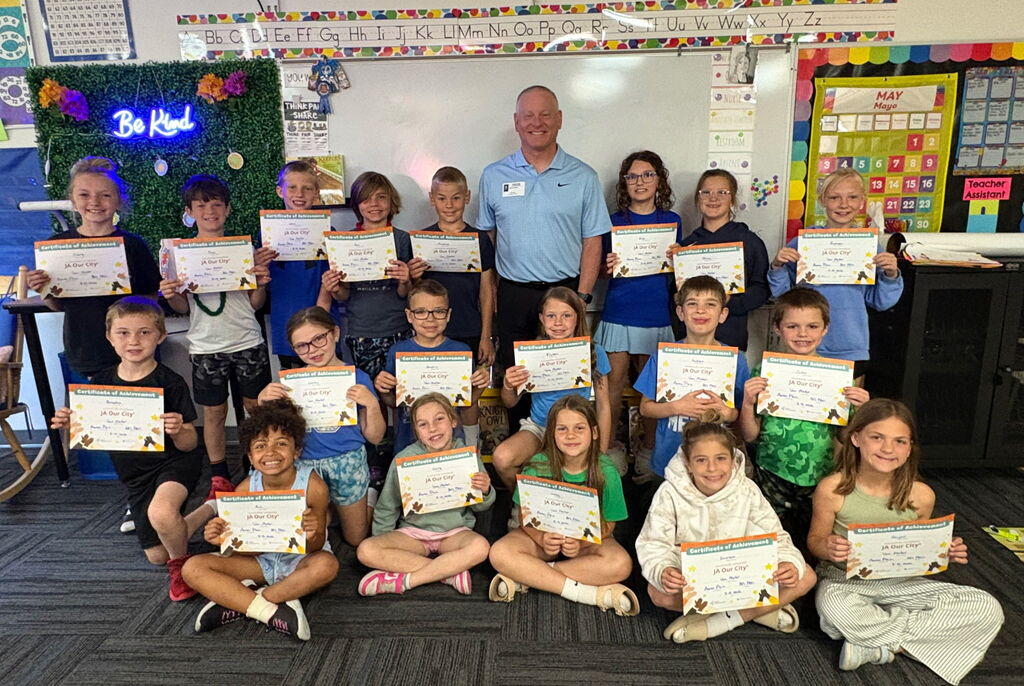 Elementary students proudly hold Junior Achievement certificates beside their teacher in a classroom group photo.