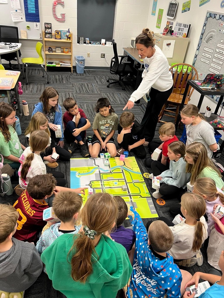 Teacher leads students seated around a city map as they discuss and place model buildings during a classroom lesson.